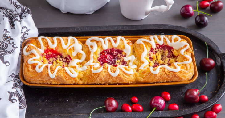 A tray of three danish pull-apart pastries, featuring golden brown flaky pastry filled with smooth cheesecake, cherries, and fresh cranberry compote, topped with honey glaze, sweet icing, and buttery streusel.