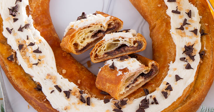 A delicious Christmas Kringle featuring a smooth cream cheese filling with rich toffee and caramel candy, topped with vanilla frosting adorned with chocolate curls and toffee bits. 